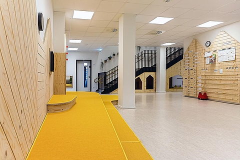 Ein Bild vom Eingangsbereich mit einem großen, gelben Podest mit mehreren Stufen auf der linken Seite. Im Hintergrund ist die schwarze Treppe mit Geländer, die ins erste Stockwerk führt. Der Raum ist hell beleuchtet und hat einen hellen Boden. 