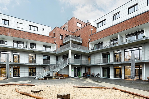  Im Vordergrund liegt ein großer Sandspielbereich mit Holzstämmen zum Balancieren. Um den Innenhof herum sind ebenerdige Eingänge mit großen Fenstern und Türen zu sehen. Auf den Balkonen und im Hof stehen Sitzmöbel.