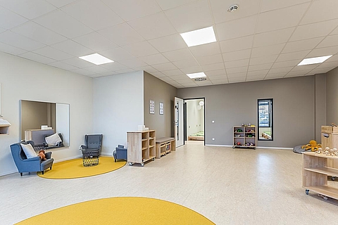 Der Rollenspielraum hat eine helle Decke und einen hellen Boden mit runden gelben Teppichen. Links befindet sich eine Sitzecke mit drei Sesseln und einem Spiegel an der Wand. Es gibt mehrere Kommoden mit Rollen, damit man sie durch den Raum schieben kann. 