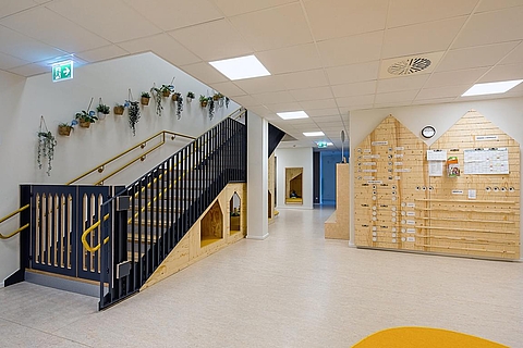 Rechts an der Wand befindet sich eine große Holzverkleidung in Hausform mit vielen Fächern. Links führt eine Treppe mit schwarzem Geländer und gelben Handläufen ins obere Stockwerk. Über der Treppe hängen mehrere kleine grüne Pflanzen in weißen Töpfen. Unter der Treppe befinden sich zwei kleine Spielecken in Form von Mini-Häusern. 