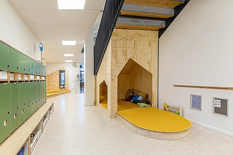 Das Bild zeigt einen hellen Flur. Links an der Wand befindet sich eine Reihe grüner Garderobenschränke. Auf der rechten Seite verläuft eine Treppe mit einem schwarzen Geländer, unter der sich eine kleine Spielecke befindet.