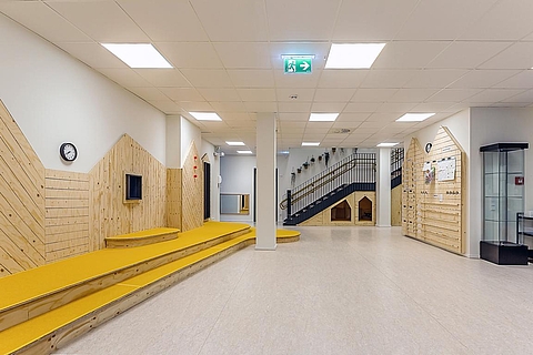 Ein heller, großer und offener Empfangsraum mir großen Deckenleuchten. Links im Bild verläuft eine hölzerne Wandverkleidung in Hausform mit integrierten Sitzstufen in Gelb. Hinten rechts führt eine breite Treppe mit schwarzem Geländer in das obere Stockwerk.