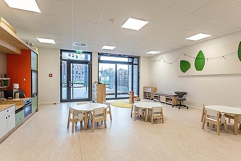 Ein heller, großer und offener Raum mit hellem Boden und heller Decke. Im Hintergrund sind große Fenster und eine Glastür. In der Mitte des Raums stehen mehrere runde Kindertische mit kleinen Holzstühlen. Links befindet sich eine kindgerechte Küchenzeile mit grün-orangefarbenen Elementen. An der rechten Wand hängen dekorative grüne Filzformen in Blattform sowie eine Lichterkette.