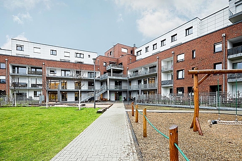 Rückseite eines modernen, mehrstöckigen Gebäudes mit einer Kombination aus rotem Klinker und weißen Fassadenelementen. Im Mittelpunkt steht ein gepflasterter Weg, der vom unteren Bildrand direkt auf das Gebäude zuführt. Links und rechts des Weges befinden sich Grünflächen und ein umzäunter Spielplatz mit Holzspielgeräten, darunter eine Schaukel und ein Balancierseil. Das Gebäude verfügt über mehrere Balkone, große Fenster und Außentreppen, die die Etagen miteinander verbinden. Im Hintergrund ist der Himmel leicht bewölkt.