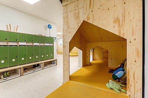 Eine nähere Aufnahme von der Spielecke unter der Treppe.  Diese ist aus Holz gebaut und hat die Form eines Häuschens. Die Spielecke ist mit gelben Matten ausgelegt. Auf den Matten liegen mehrere Kuscheltiere. Links neben der Spielecke befinden sich die grünen Garderobenschränke. 